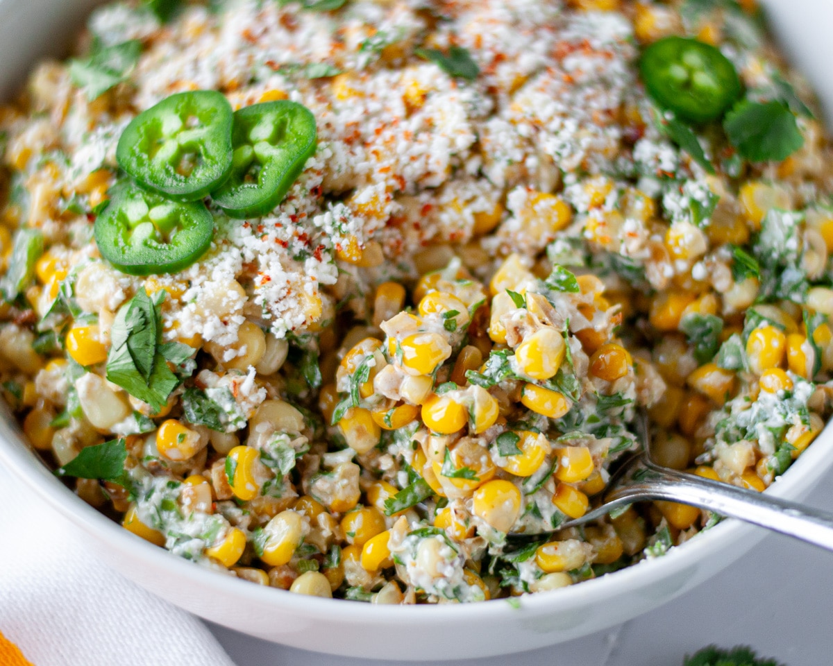 A serving spoon with a scoop of Mexican street corn salad sitting in the serving bowl with extra cotija cheese, tajin, jalapeño slices, and cilantro on top.