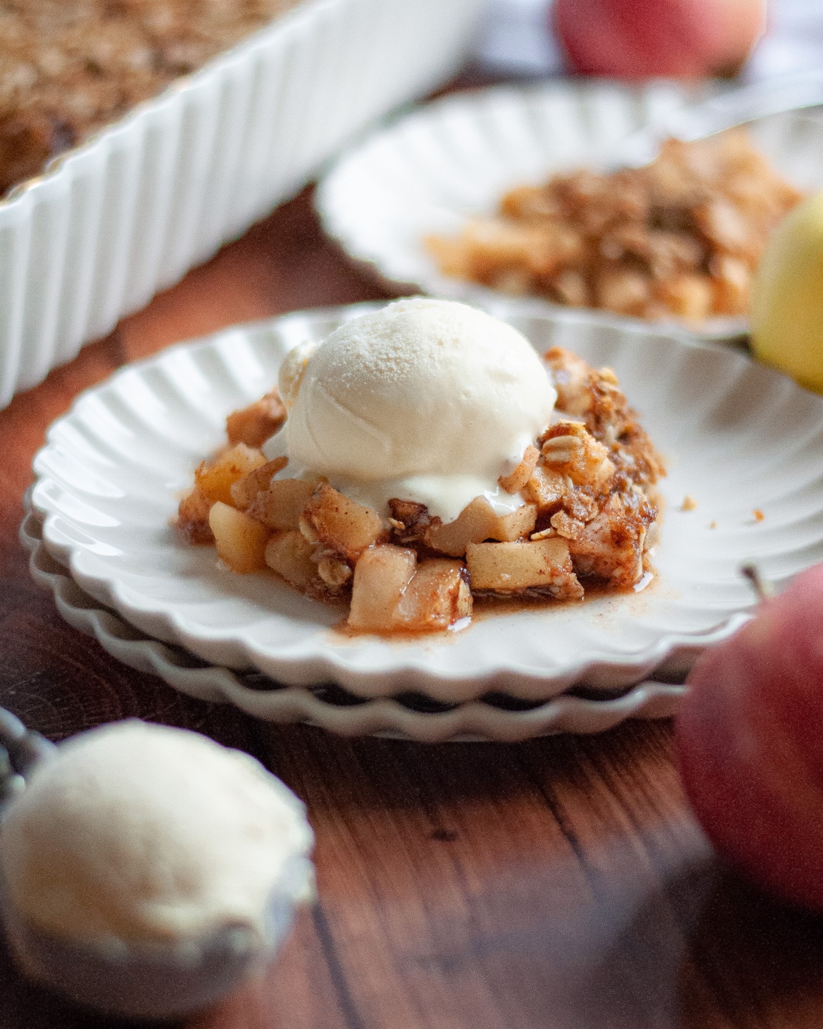 Close up of a serving of apple crisp on a delicate white plate topped with a scoop of vanilla ice cream, with another serving of apple crisp in the background next to the 9x13 pan filled with the rest of the crisp.