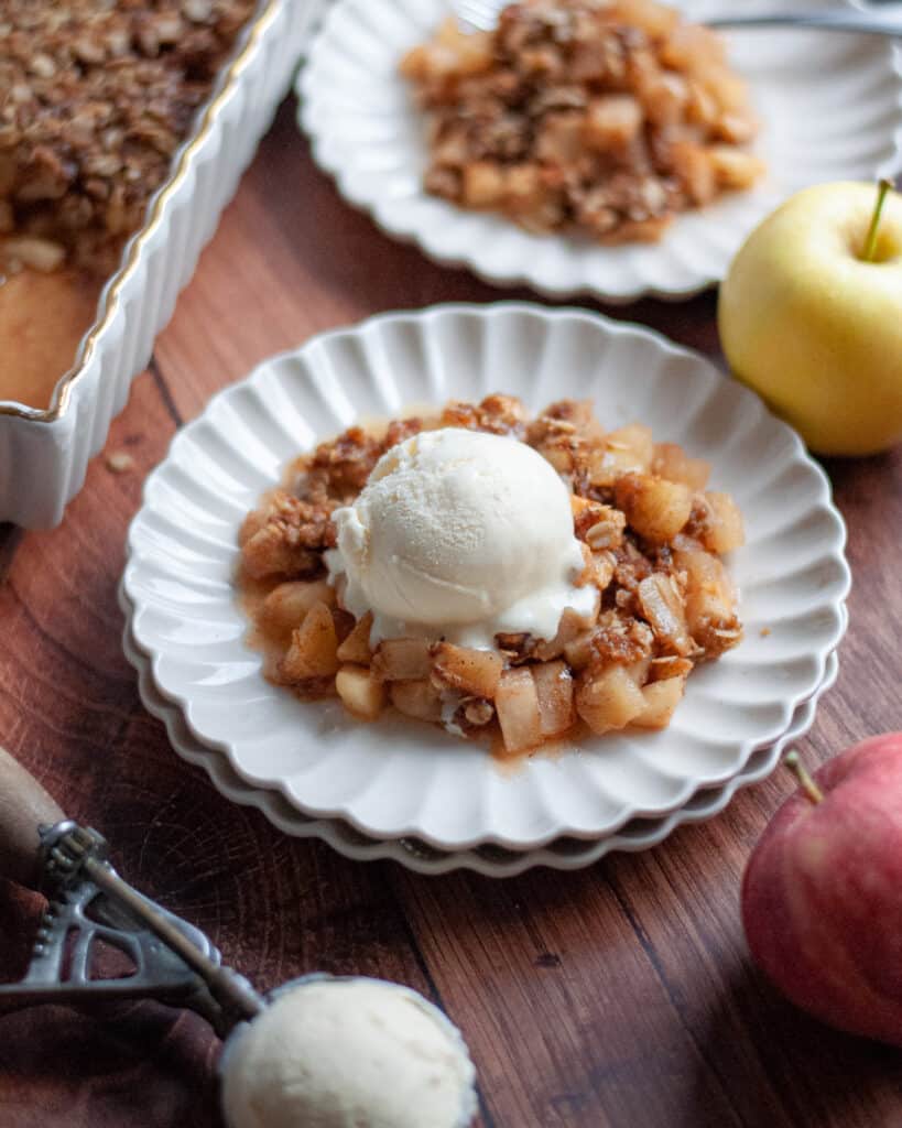 Close up of a serving of apple crisp on a delicate white plate topped with a scoop of vanilla ice cream, with another serving of apple crisp in the background next to the 9x13 pan filled with the rest of the crisp.