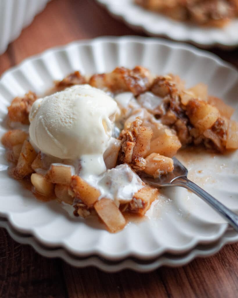 Close up of a serving of apple crisp on a delicate white plate topped with a scoop of vanilla ice cream with a fork digging into the apple crisp, ready to take a bite of this fall dessert.