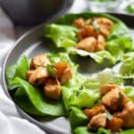 A dark grey plate filled with mango chicken lettuce wraps garnished with cilantro with a white linen on the side of the plate, and bowls of ingredients and additional filling in the background.
