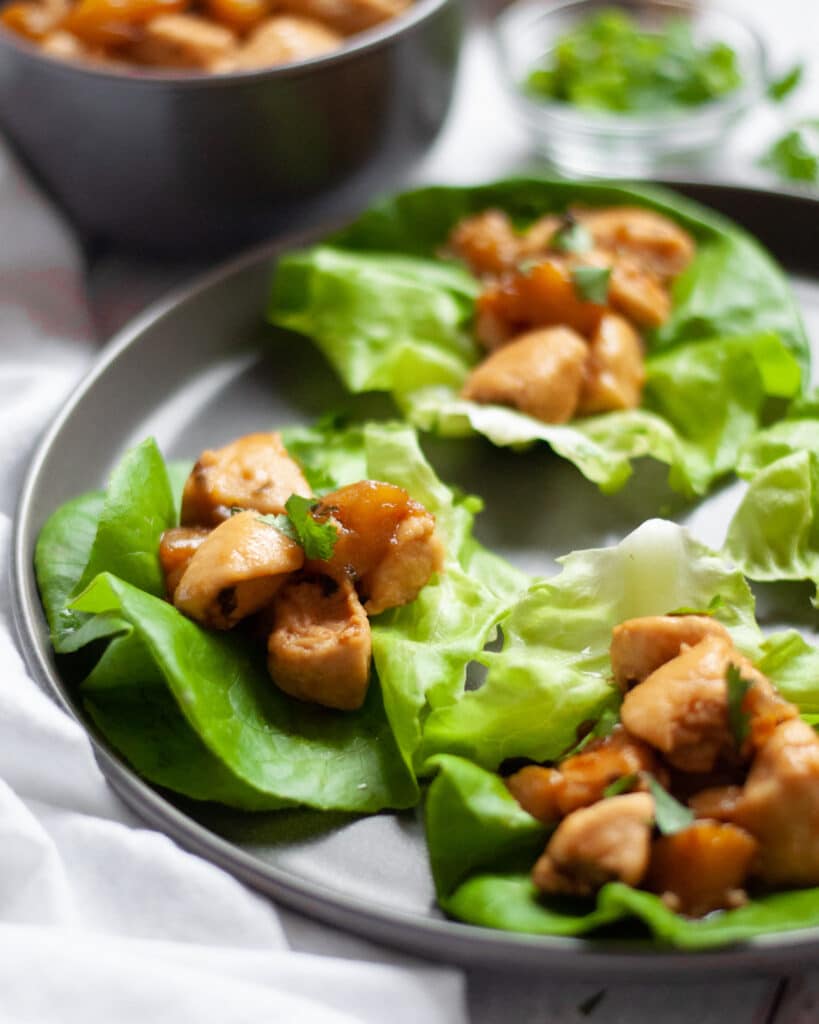 A dark grey plate filled with mango chicken lettuce wraps garnished with cilantro with a white linen on the side of the plate, and bowls of ingredients and additional filling in the background.