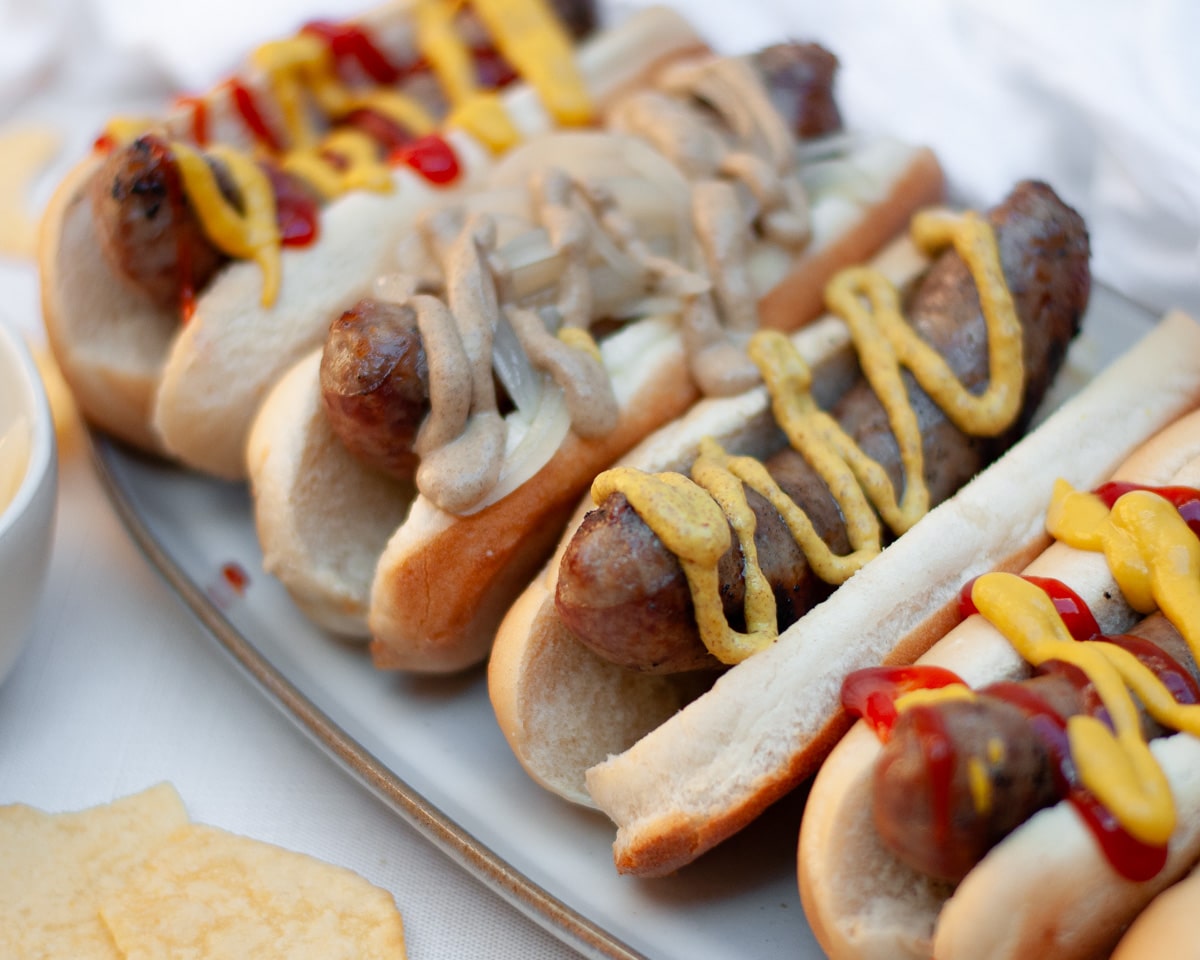 Close up of a serving tray covered with beer brats on buns and topped with a variety of toppings like onions, mustard, and ketchup.