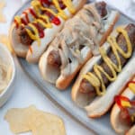 Close up of a serving tray covered with beer brats on buns and topped with a variety of toppings like onions, mustard, and ketchup.