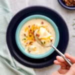 A hand holding a spoonful of cheesy potato soup above a serving bowl of soup.