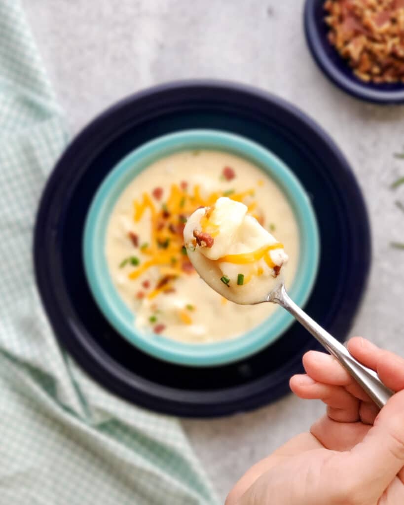 A hand holding a spoonful of cheesy potato soup above a serving bowl of soup.