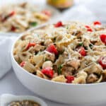 A serving dish filled high with chicken and pesto pasta salad with a serving spoon in it, and a white linen, plate of pasta, and bowl of pesto around it.