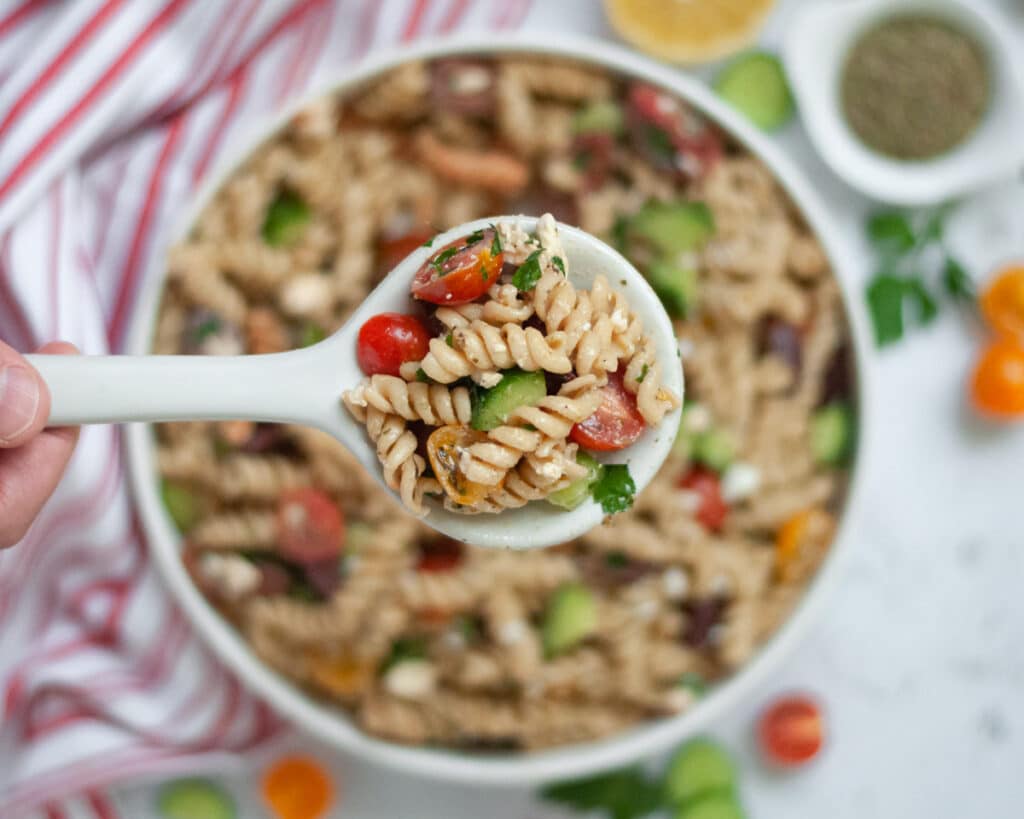 Top down shot of vegetarian Mediterranean Pasta Salad in a white serving bowl with a spoonful held up closer to the camera.