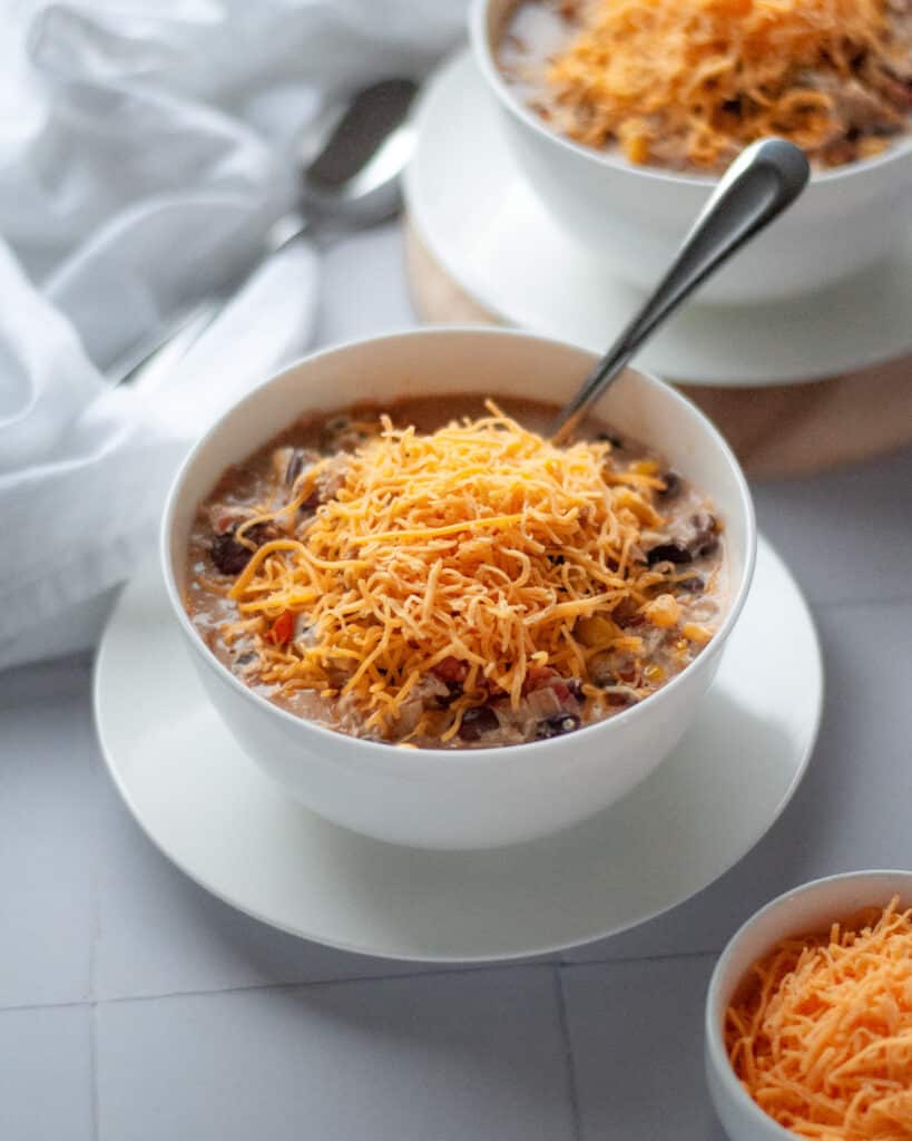 A bowl of white chicken chili topped with shredded cheese with a spoon in the bowl with an additional bowl of chili peaking into the background of the image.