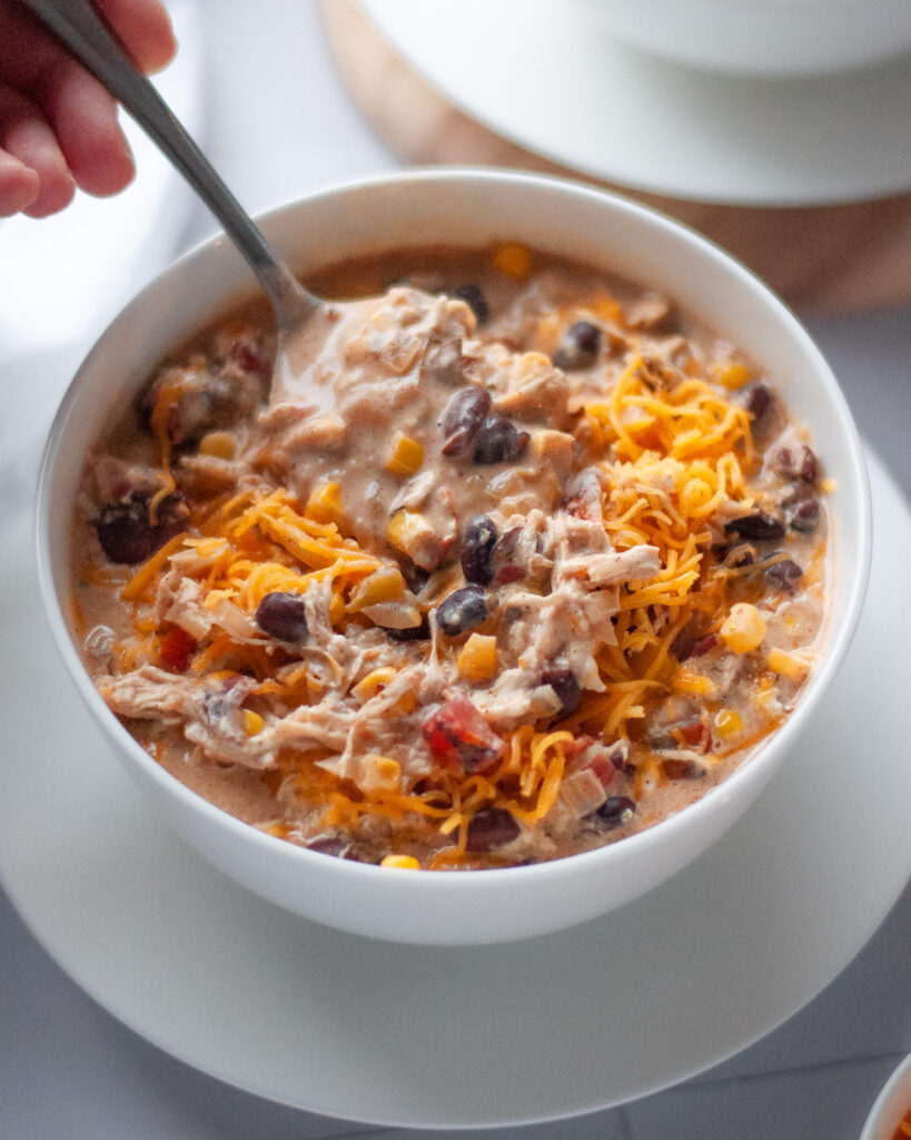 A bowl of white chicken chili with a hand taking a spoonful of chili to eat.