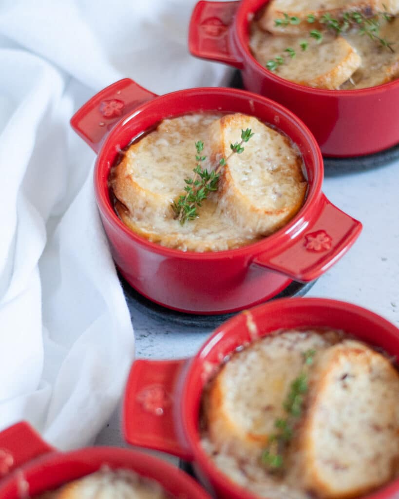 Close up of a red ramekin of French onion soup, topped with baguette slices, cheese, and fresh thyme sprigs with additional crocks of French onion soup surround the main dish.