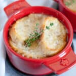 Close up of a red ramekin of French onion soup, topped with baguette slices, cheese, and fresh thyme sprigs with additional crocks of French onion soup surround the main dish.