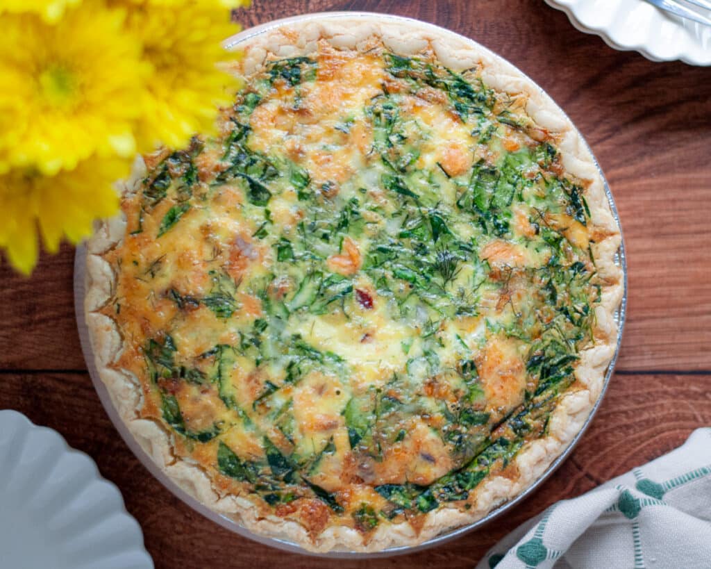 Top down view of a full smoked salmon and spinach quiche with serving plates, a linen, and yellow flowers also peaking into the frame.