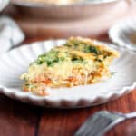 A slice of smoked salmon and spinach quiche on a serving plate is the focus with the pie pan of spinach salmon quiche blurry in the background.