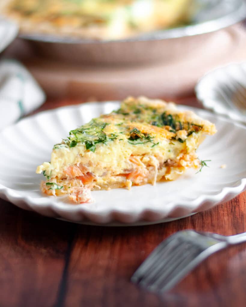 A slice of smoked salmon and spinach quiche on a serving plate is the focus with the pie pan of spinach salmon quiche blurry in the background.