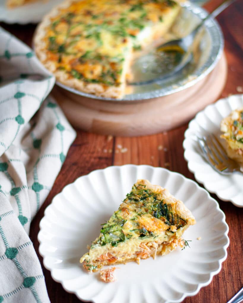 A slice of smoked salmon and spinach quiche on a serving plate with additional serving plates and the pie pan of spinach salmon quiche are in the background.