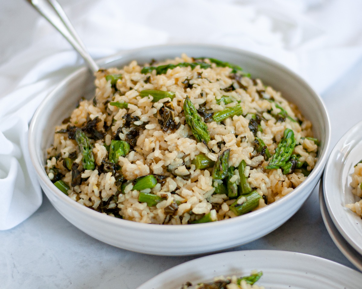 Close-up view of a white bowl of Summer Vegetable Risotto with asparagus and kale with a serving spoon in the bowl.