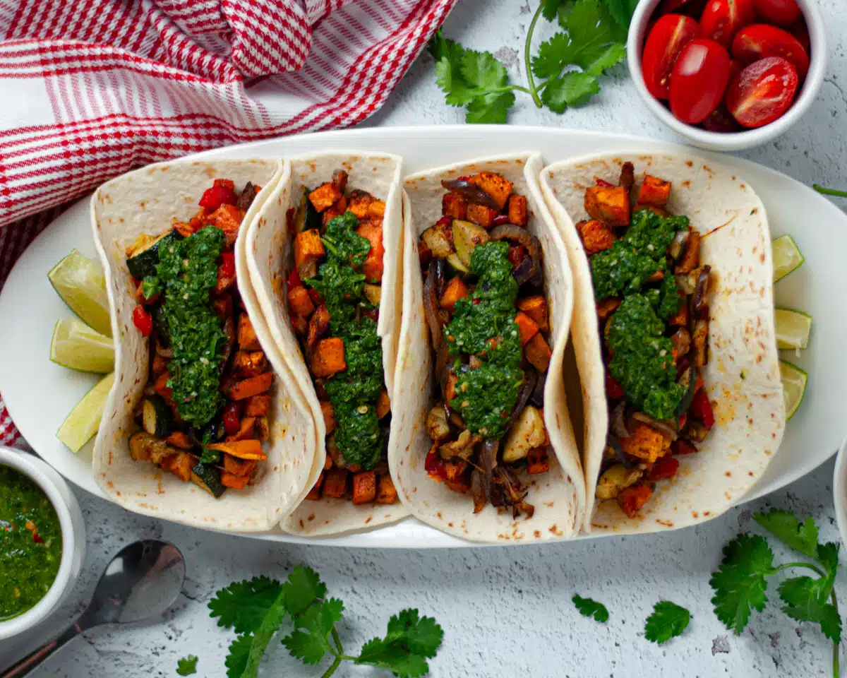 A white plate holds four soft-shell tacos filled with roasted vegetables and topped with green sauce, garnished with lime wedges. A bowl of cherry tomatoes, a small dish of grated cheese, a spoon of green sauce, and a red and white checkered cloth are beside the plate.