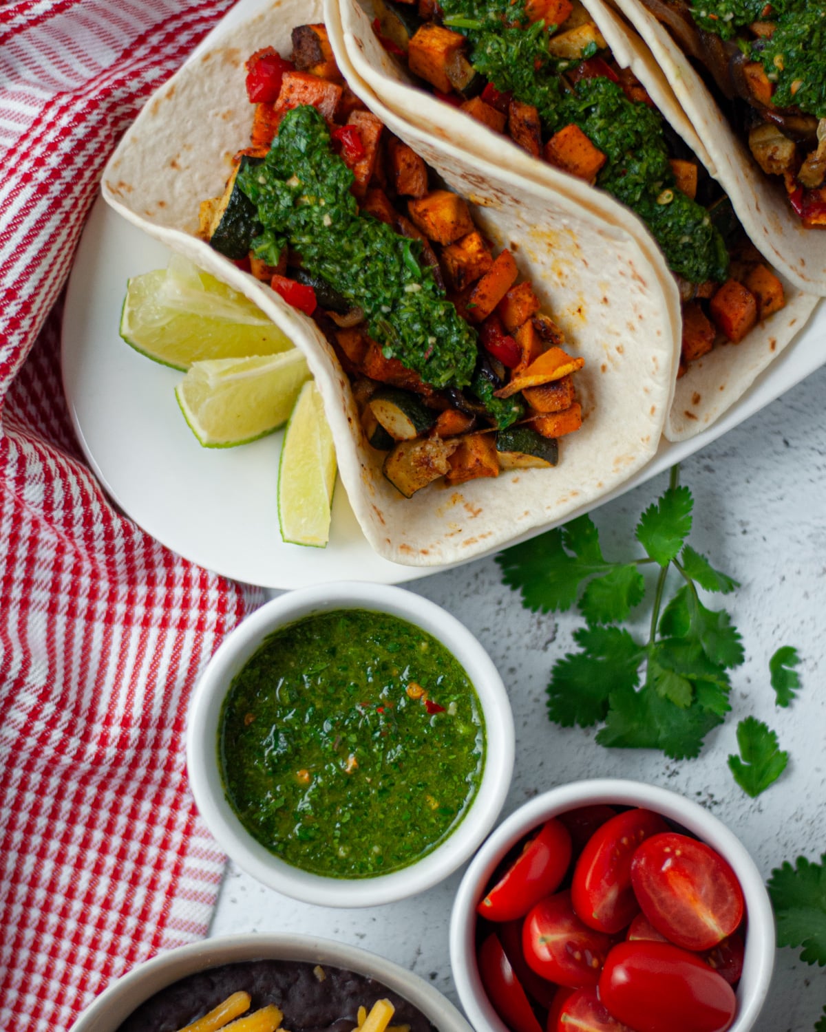 Top down view of three vegetable tacos served with a side of lime wedges, chimichurri sauce, tomatoes, and a side of refried beans.