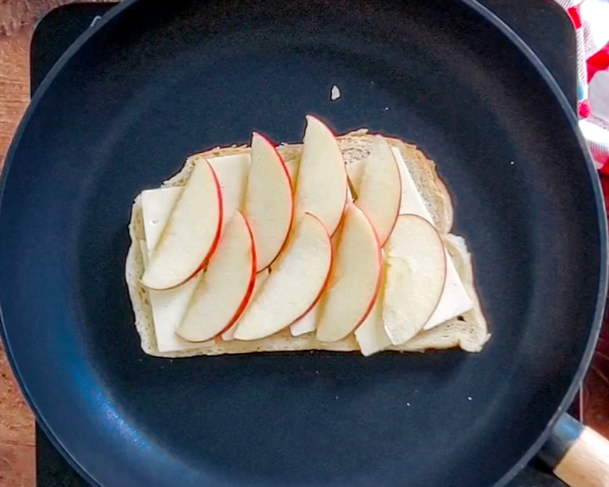 Process shot showing one slice of bread butter side down in the skillet with a layer of apple slices over the first layer of cheese slices.