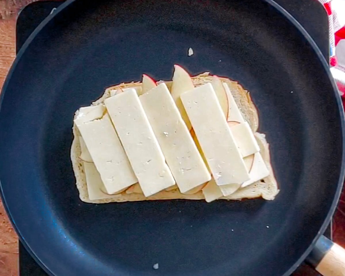 Process shot showing one slice of bread butter side down in the skillet with a a second layer of cheese over the layer of apple slices that's over the first layer of cheese slices.