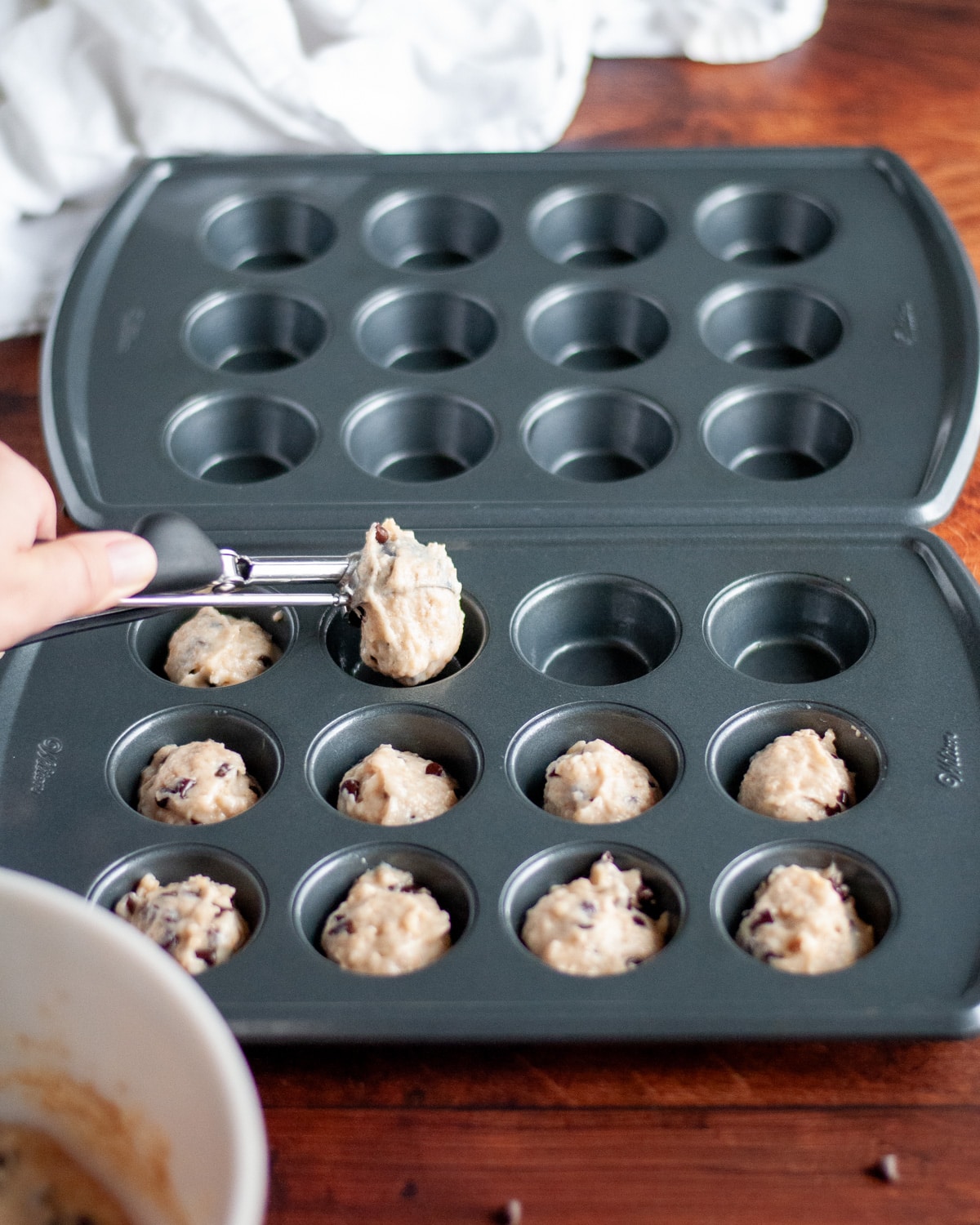 Process shot showing the chocolate chip muffin batter being added to a mini muffin tin using a small cookie scoop.