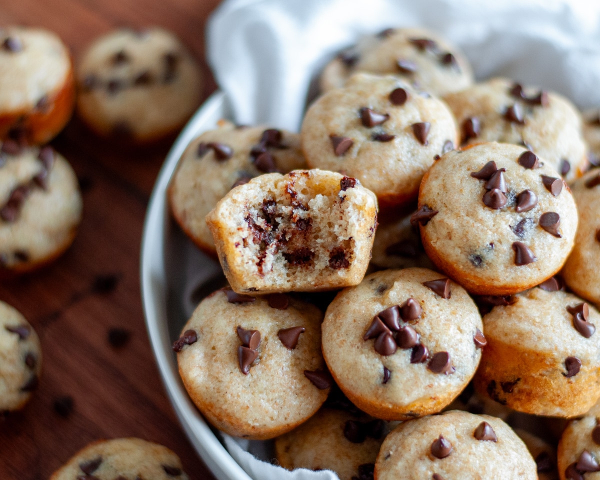 Close up of a bowl filled with chocolate chip mini muffins, with one muffin having a bite out of it to see the inside.