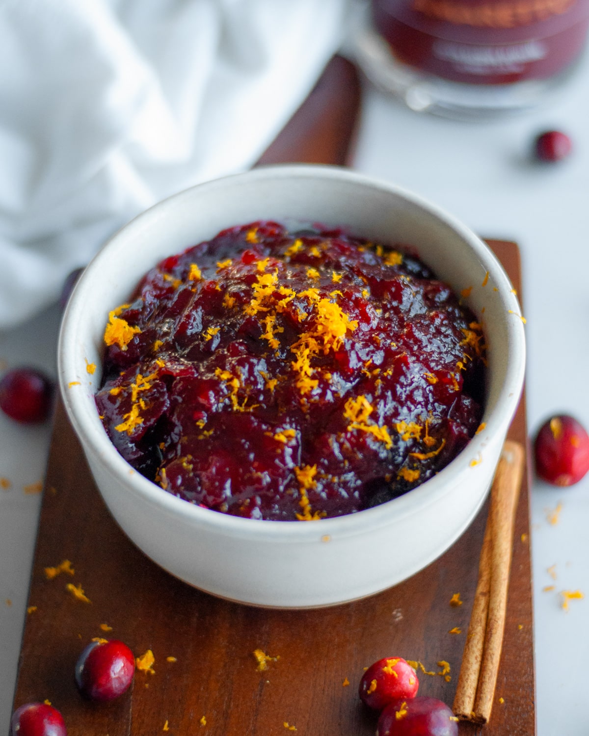 Close up of a bowl of homemade cranberry sauce with orange and amaretto topped with orange zest on a wood board with fresh cranberries, a cinnamon stick, and a white linen around it.