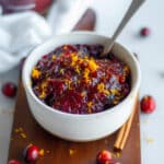A bowl of homemade cranberry sauce with orange and amaretto topped with orange zest on a wood board with fresh cranberries, a cinnamon stick, and a white linen around it.