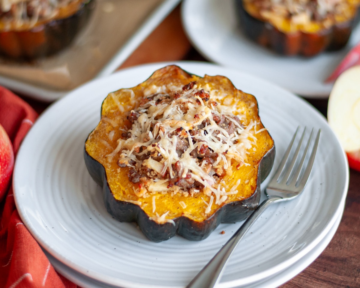 A sausage and apple stuffed acorn squash half on a serving plate with a fork. Apples, apple slices, and addition stuffed squash halves surround the plate.