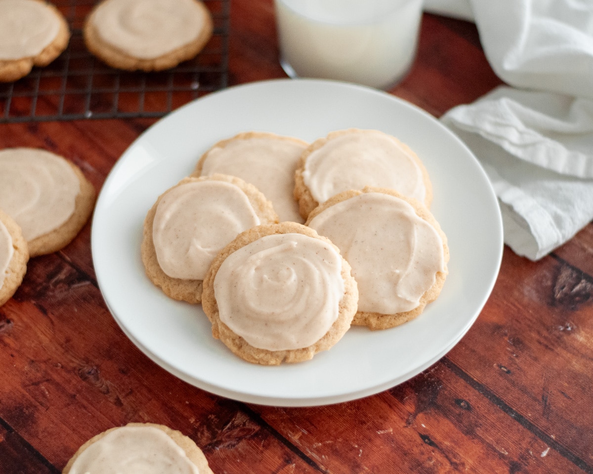 A white plate full of cardamom cookies with cardamom frosting, with a white linen, glass of milk, and additional cookies around it.