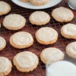 Cardamom frosted cookies on a wire rack with glasses of milk and plate of cookies in the background.