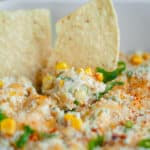 Close up of a chip scooping up hot Mexican street corn dip in a white baking dish.