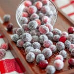 A fancy lowball glass spilling sugared cranberries out onto a wooden tray.
