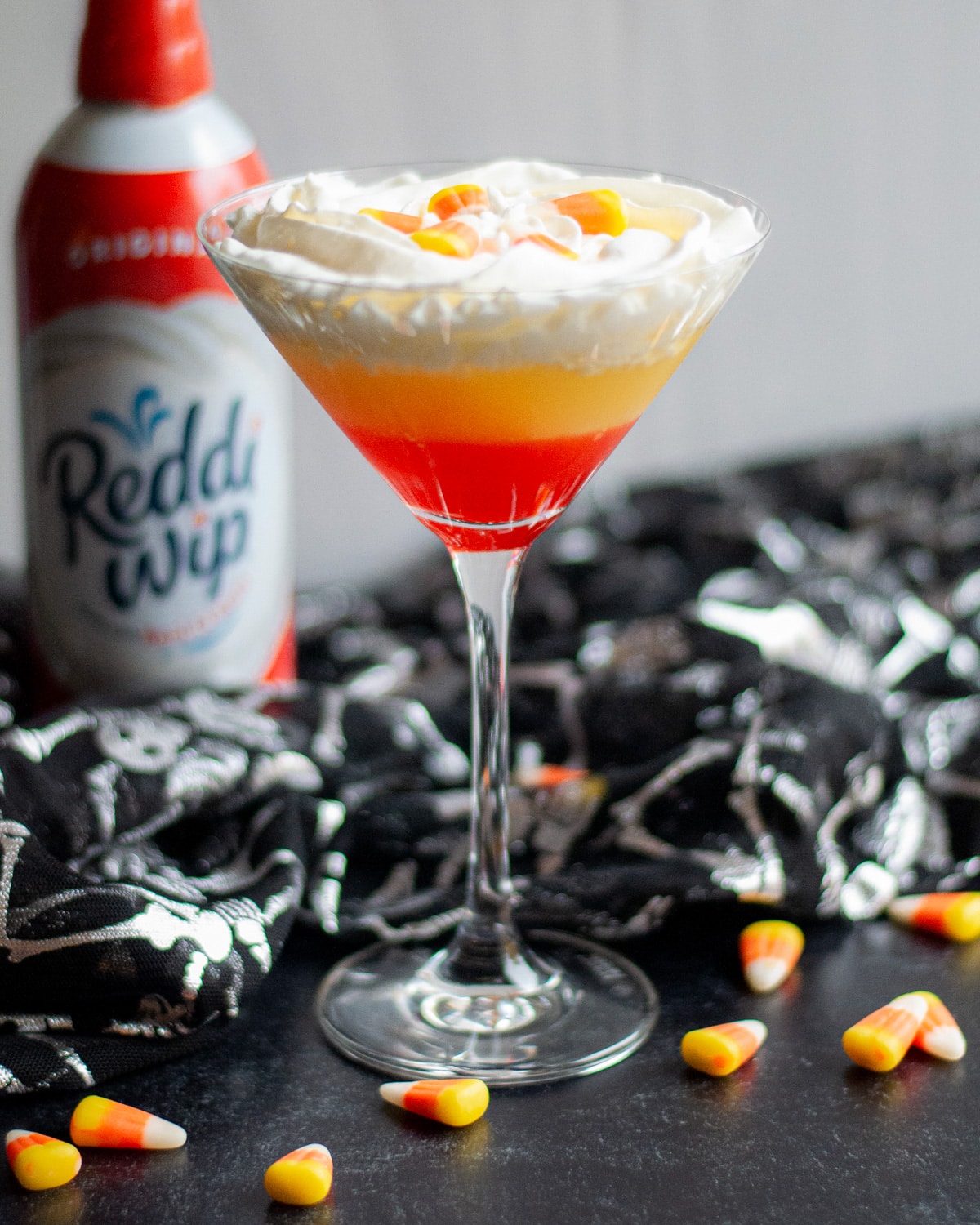 Close up of a martini glass with a layered candy corn martini in it, surrounded by candy corn pieces, a black and silver skeleton cloth, and a can of squirt whipped cream.