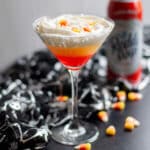 Close up of a martini glass with a layered candy corn martini in it, surrounded by candy corn pieces, a black and silver skeleton cloth, and a can of squirt whipped cream.