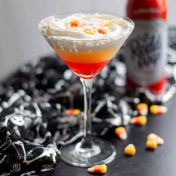 Close up of a martini glass with a layered candy corn martini in it, surrounded by candy corn pieces, a black and silver skeleton cloth, and a can of squirt whipped cream.