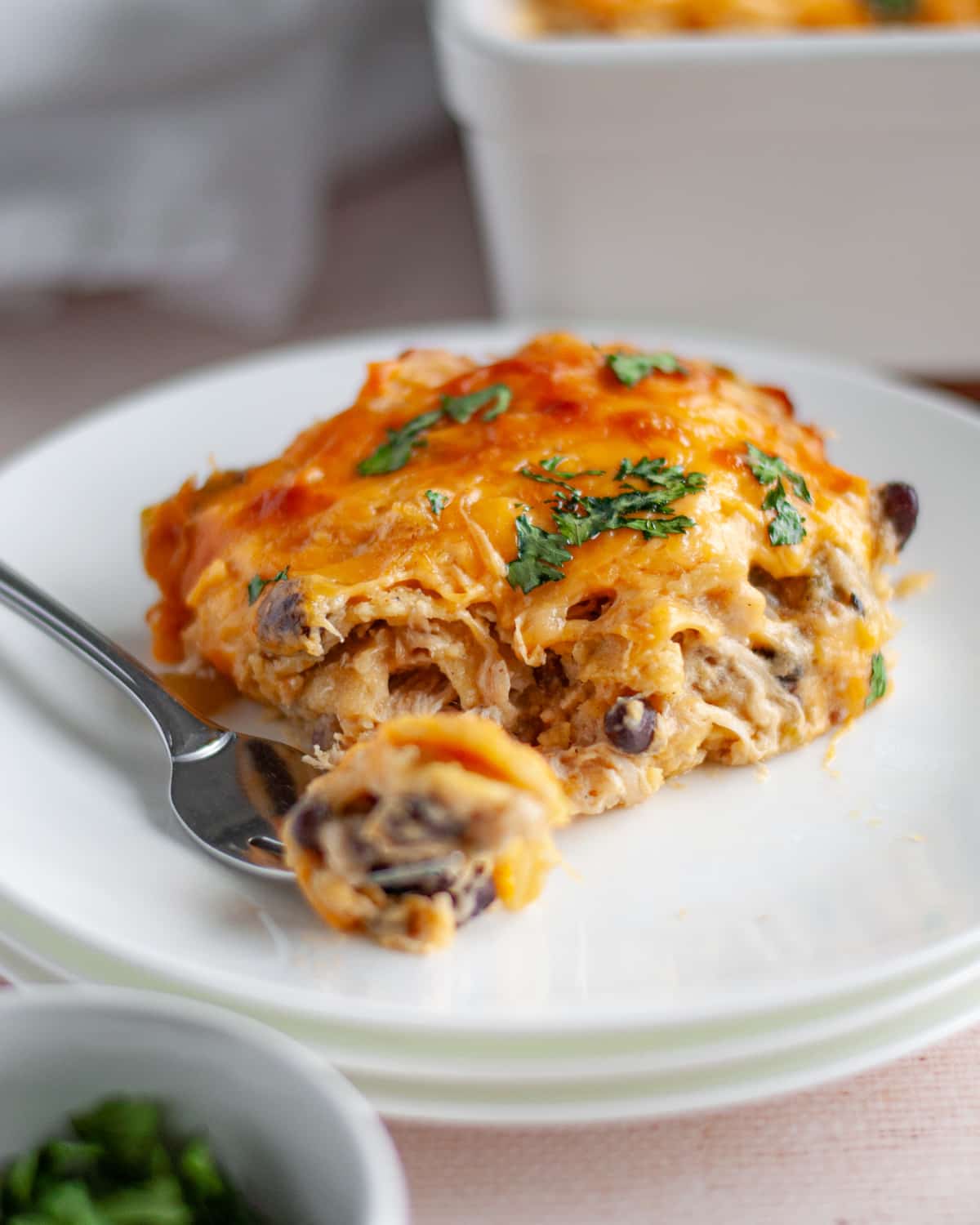 Close up of a slice of chicken enchilada bake on a white serving dish with a bite of the casserole sitting on a fork on the plate.