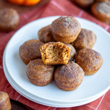 A plate full of pumpkin mini muffins with one muffin having a bite out of it; the plate sits on an orange linen with more muffins and a few pumpkins scattered around it.