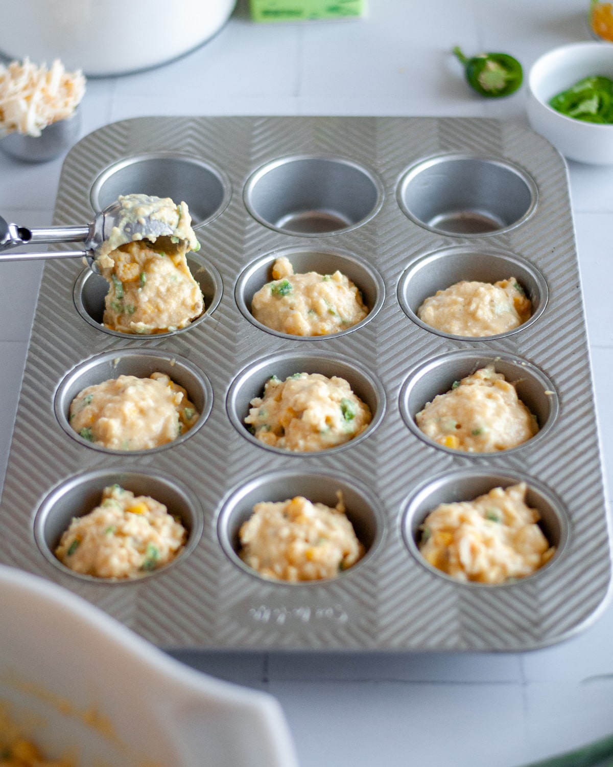 Process shot showing the batter being spooned into the greased muffin tins with a large cookie scoop.