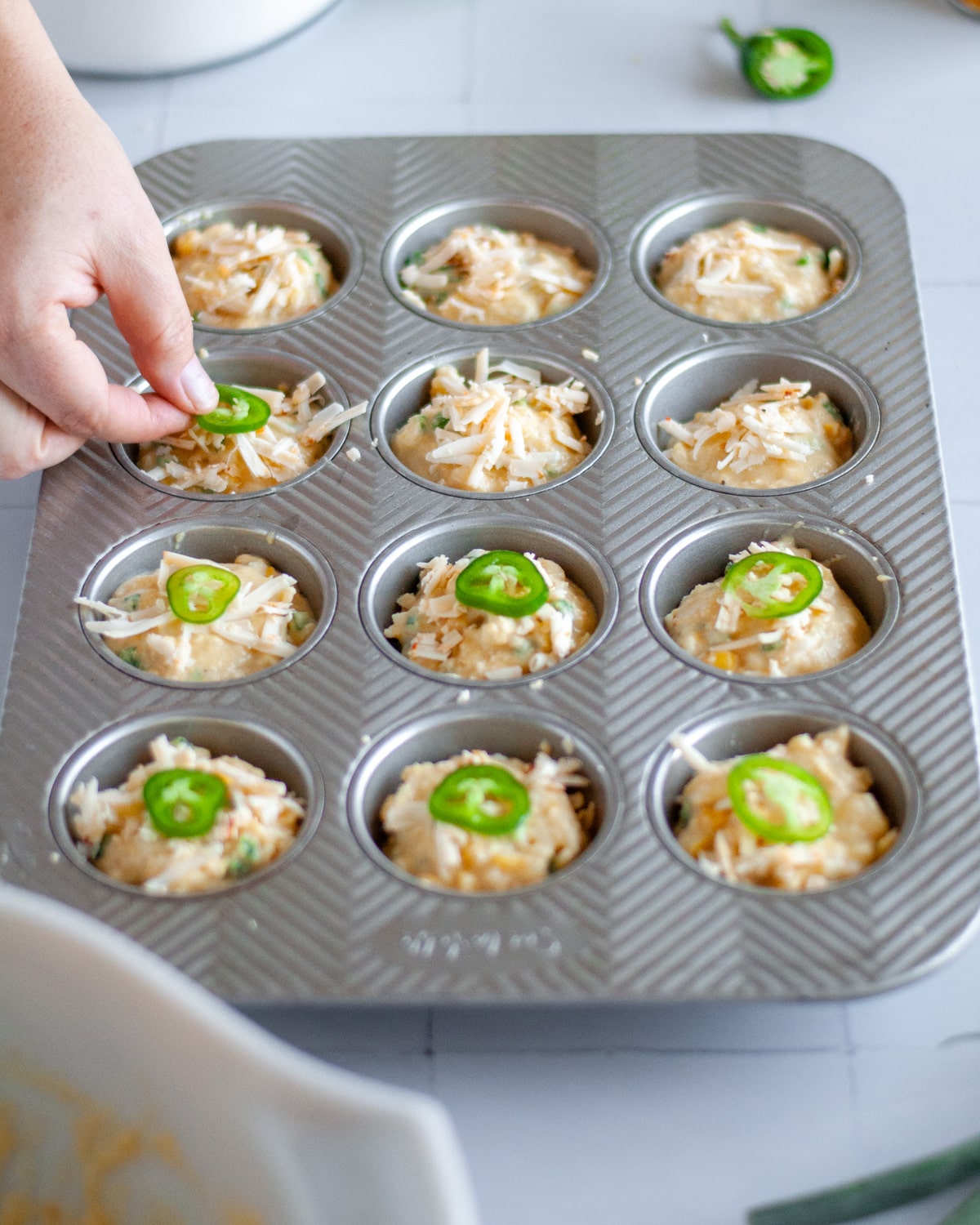 Process shot showing jalapeno pieces being set on top of the cornbread muffin batter and extra shredded cheese.