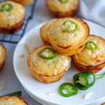 Close up of a plate of jalapeno and cheese cornbread muffins with additional muffins, jalapeno pieces, shredded cheese, and a white linen around it.