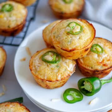 Close up of a plate of jalapeno and cheese cornbread muffins with additional muffins, jalapeno pieces, shredded cheese, and a white linen around it.
