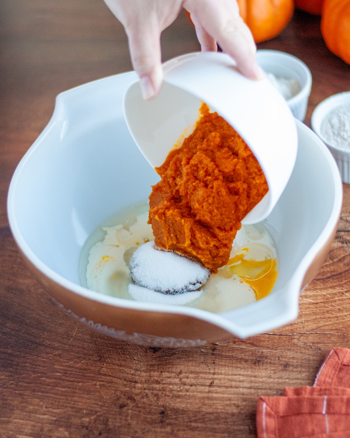 Process shot showing the pumpkin puree being added to a large mixing bowl with the rest of the wet ingredients and sugars.