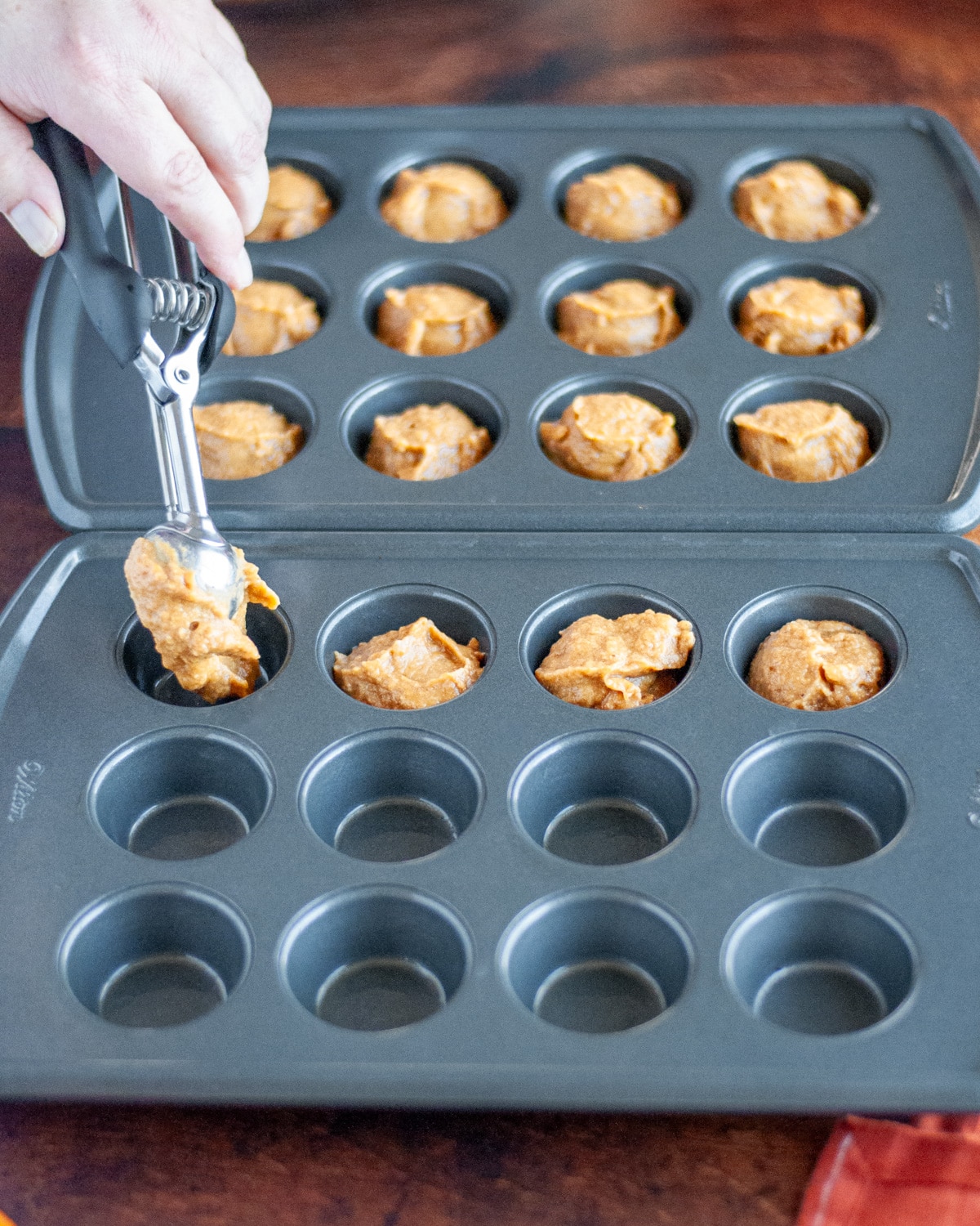 Process shot showing the batter being scooped into the greased mini muffin tin with a small cookie scoop.