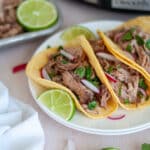 A white plate with three carnitas tacos topped with fresh cilantro, radishes, and served with lime wedges with the pan of carnitas and the crockpot in the background.