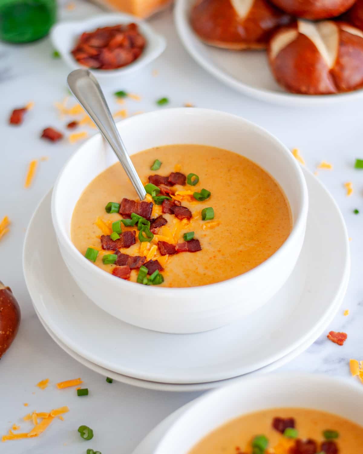 A bowl of beer and cheese soup with a spoon in it and garnished with extra shredded cheese, bacon pieces, and green onion.