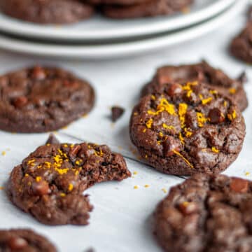Close up of a two orange chocolate cookies, one with a bite out of it, topped with orange zest surrounded by more cookies, chocolate chips, and orange zest.