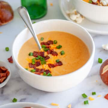 Feature image for our beer and cheese soup recipe showing a bowl of soup with a spoon in it and garnished with extra shredded cheese, bacon pieces, and green onion.