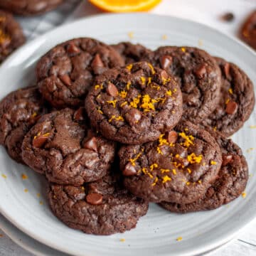 Feature image for our chocolate orange cookies recipe showing a serving plate stacked with a pile of cookies with orange zest on top of a few of them.